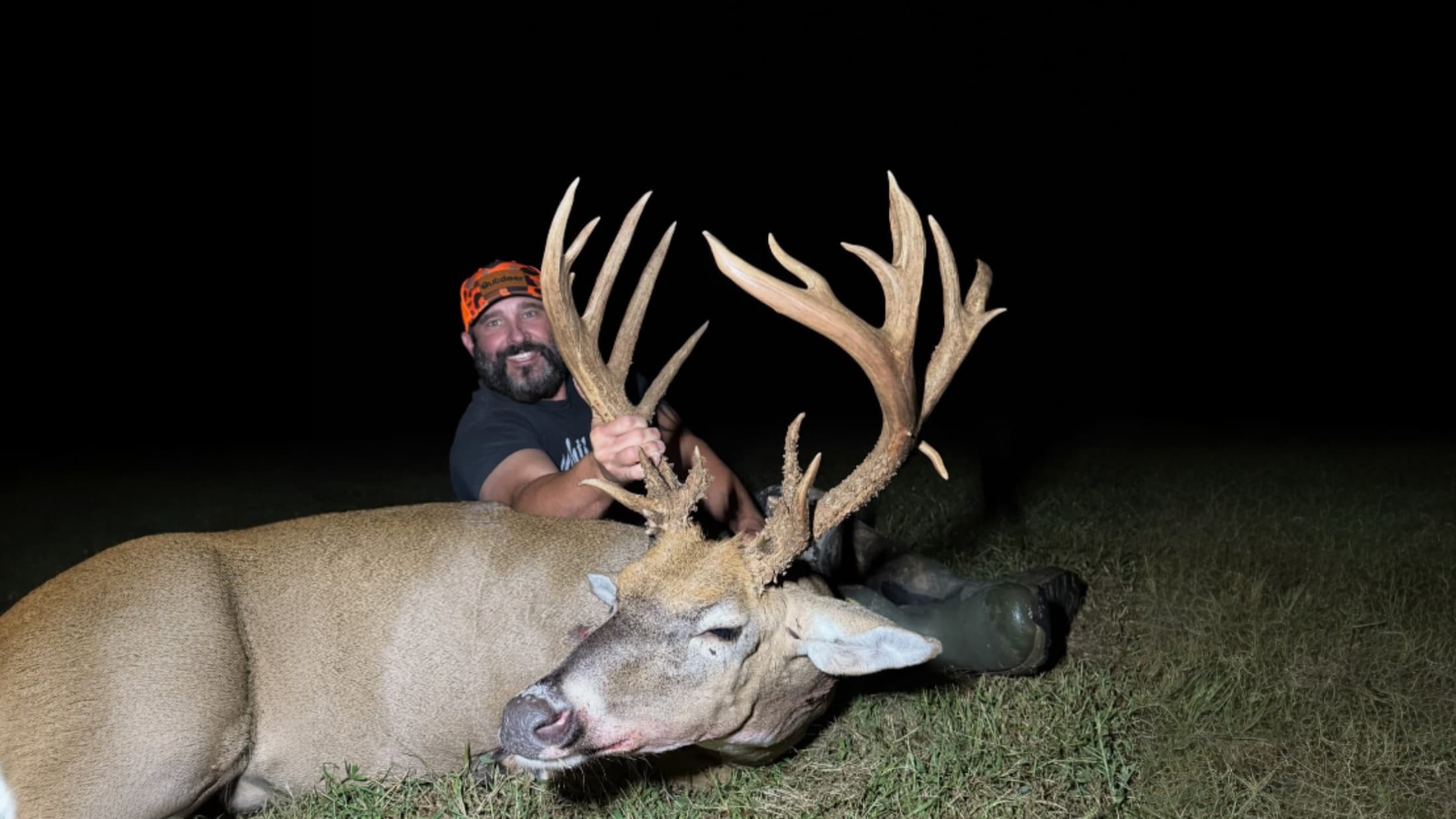 Man smiling, holding antlers of a large buck. The deer is lying on grass. Man wears an orange hunting cap, dark shirt, bea...