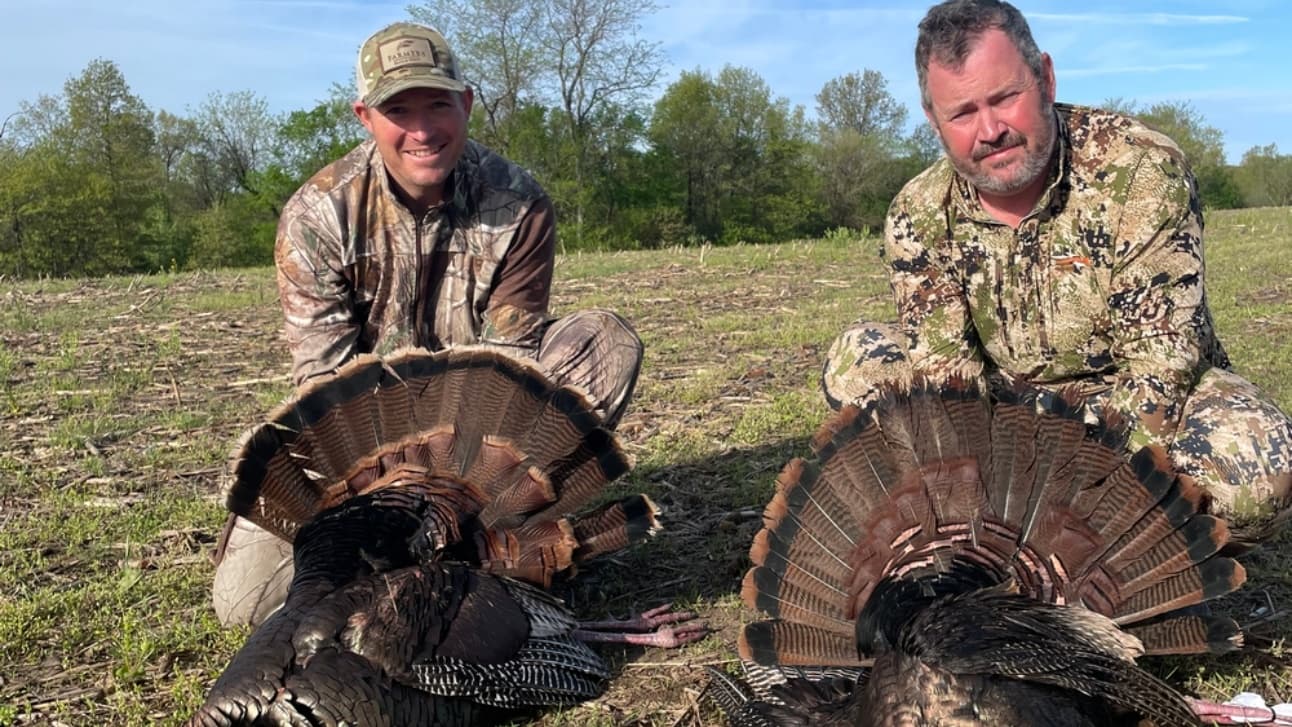 Two hunters in camouflage pose with two fan-tailed wild turkeys in a grassy field.