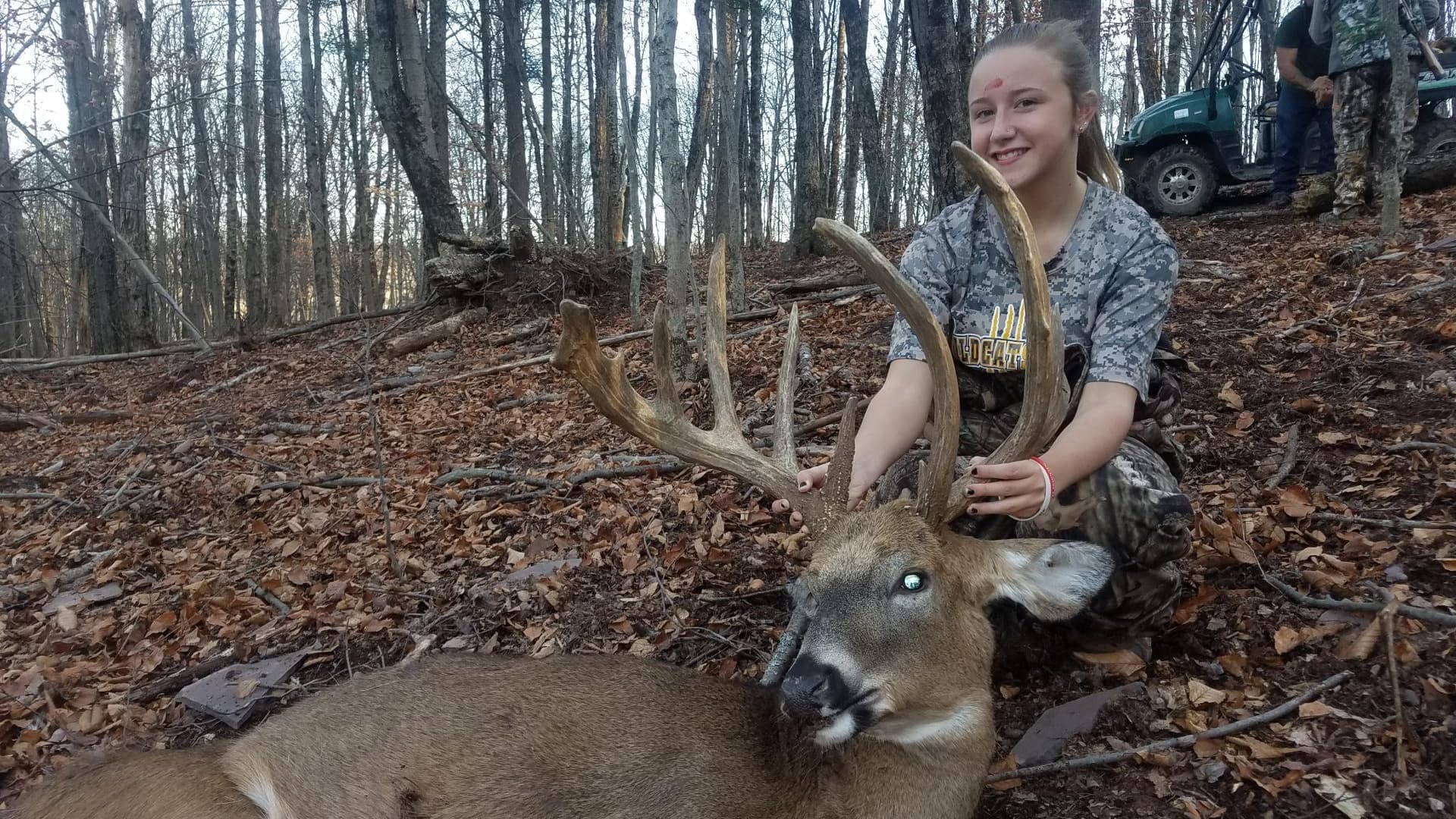 A girl kneels beside a large buck with impressive antlers. The deer lies on forest floor with fall leaves. A utility veh...
