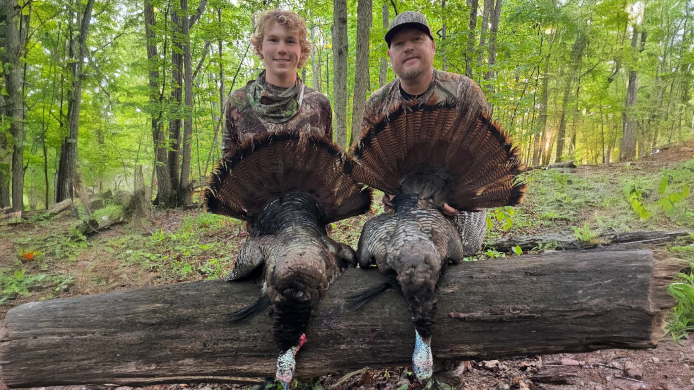 Two hunters pose with two turkeys, each holding a fan of feathers. They sit on a log in a forest setting. Both wear camo.