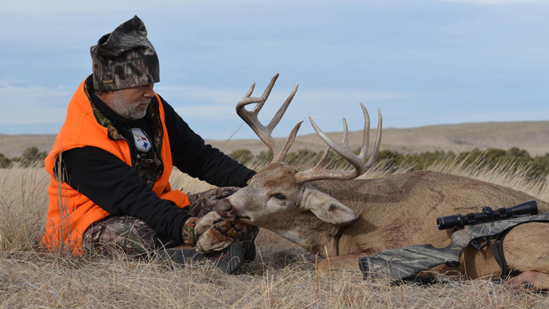Hunter in orange vest kneels by a dead deer with large antlers. Rifle rests beside the deer in a grassy field.