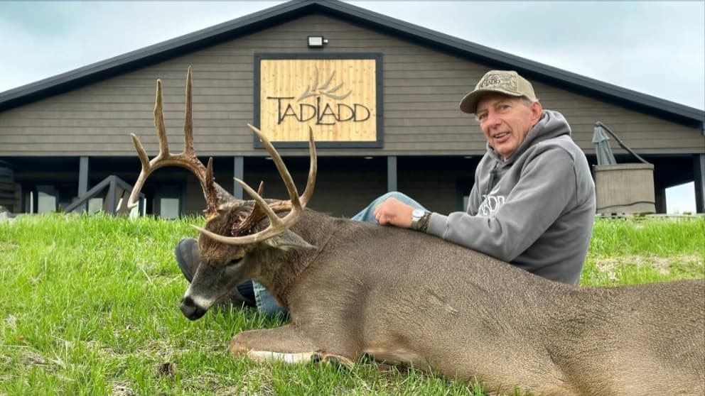 Man sitting next to a large buck with impressive antlers on green grass. Building with "TADADD" sign in the background.