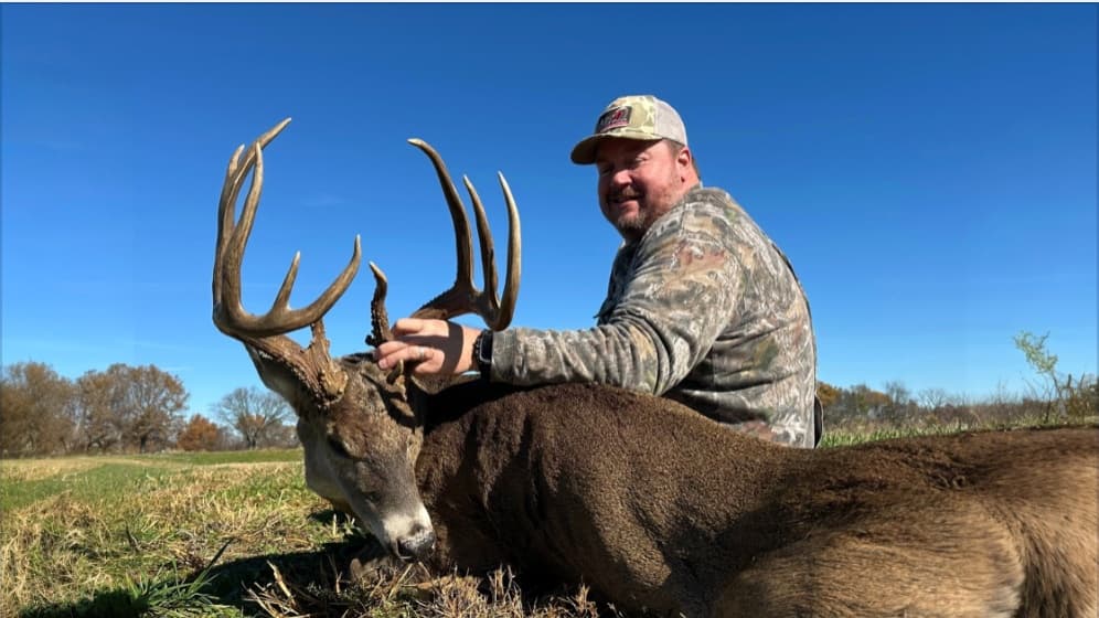Man in camo hat and shirt, posing with a large buck. Buck has large antlers. Blue sky and grassy field in the background.