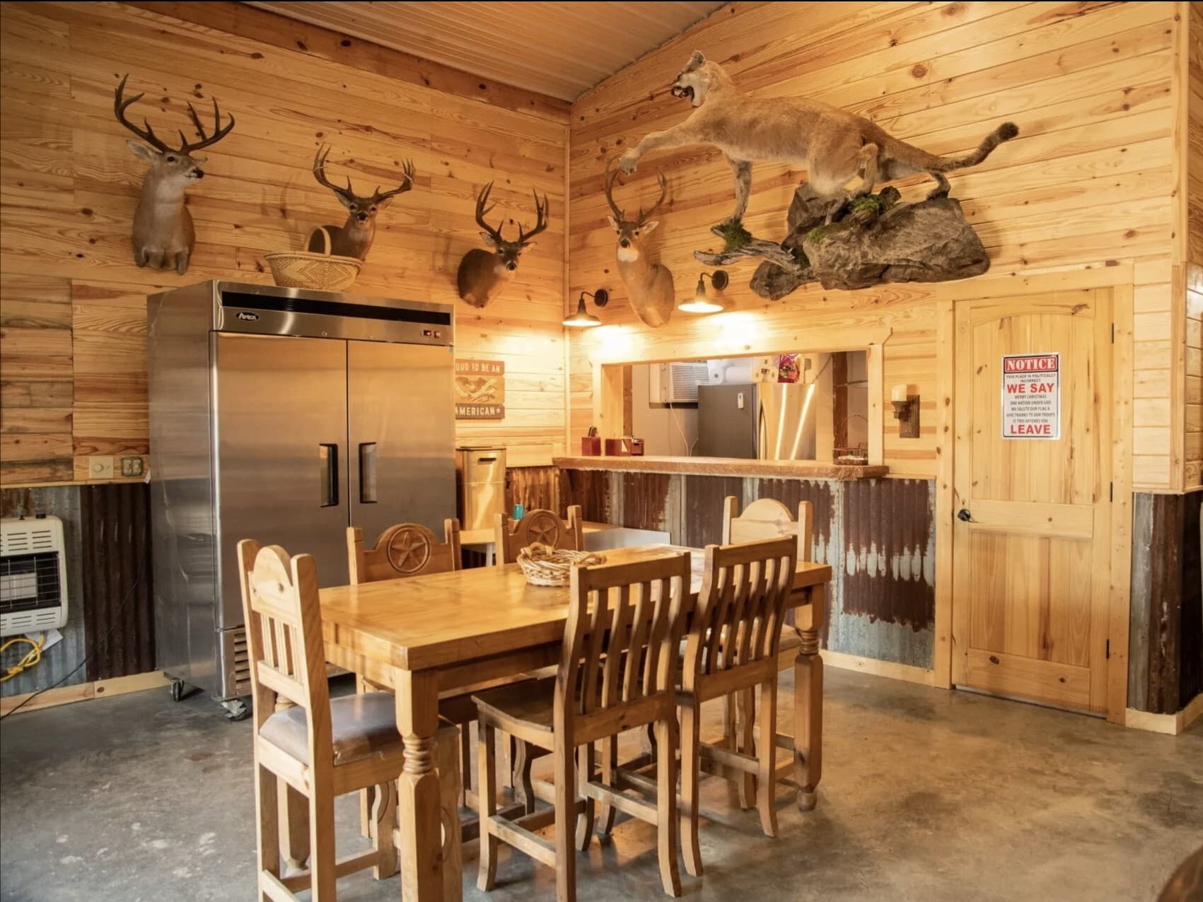 Taxidermied mountain lion above deer mounts in a rustic room with a stainless steel fridge and wooden table.