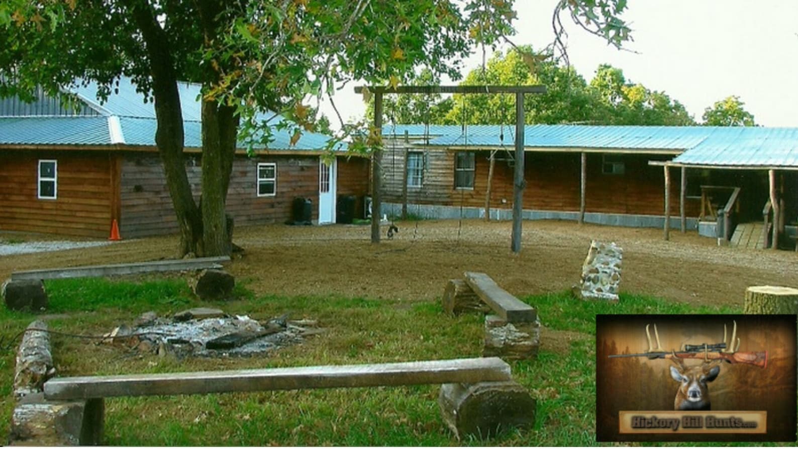 Cabin with green metal roof, a swing set, fire pit, and rustic benches in a grassy area. Hickory Hill Hunts logo is in the...