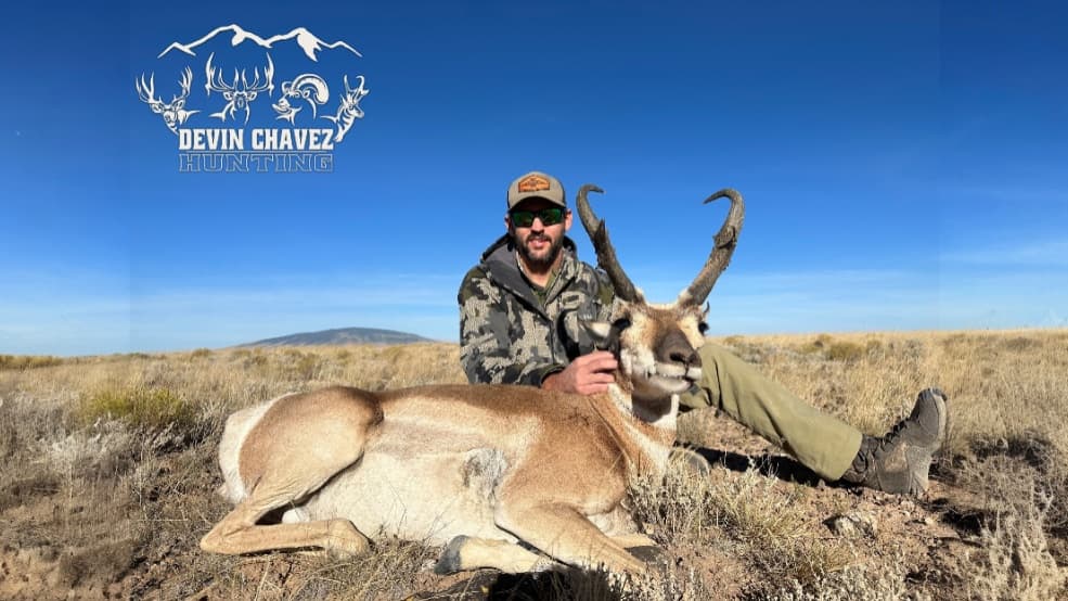 A man in camo and sunglasses with a harvested pronghorn. The animal is lying on the ground. Clear blue sky. Logo at the top.