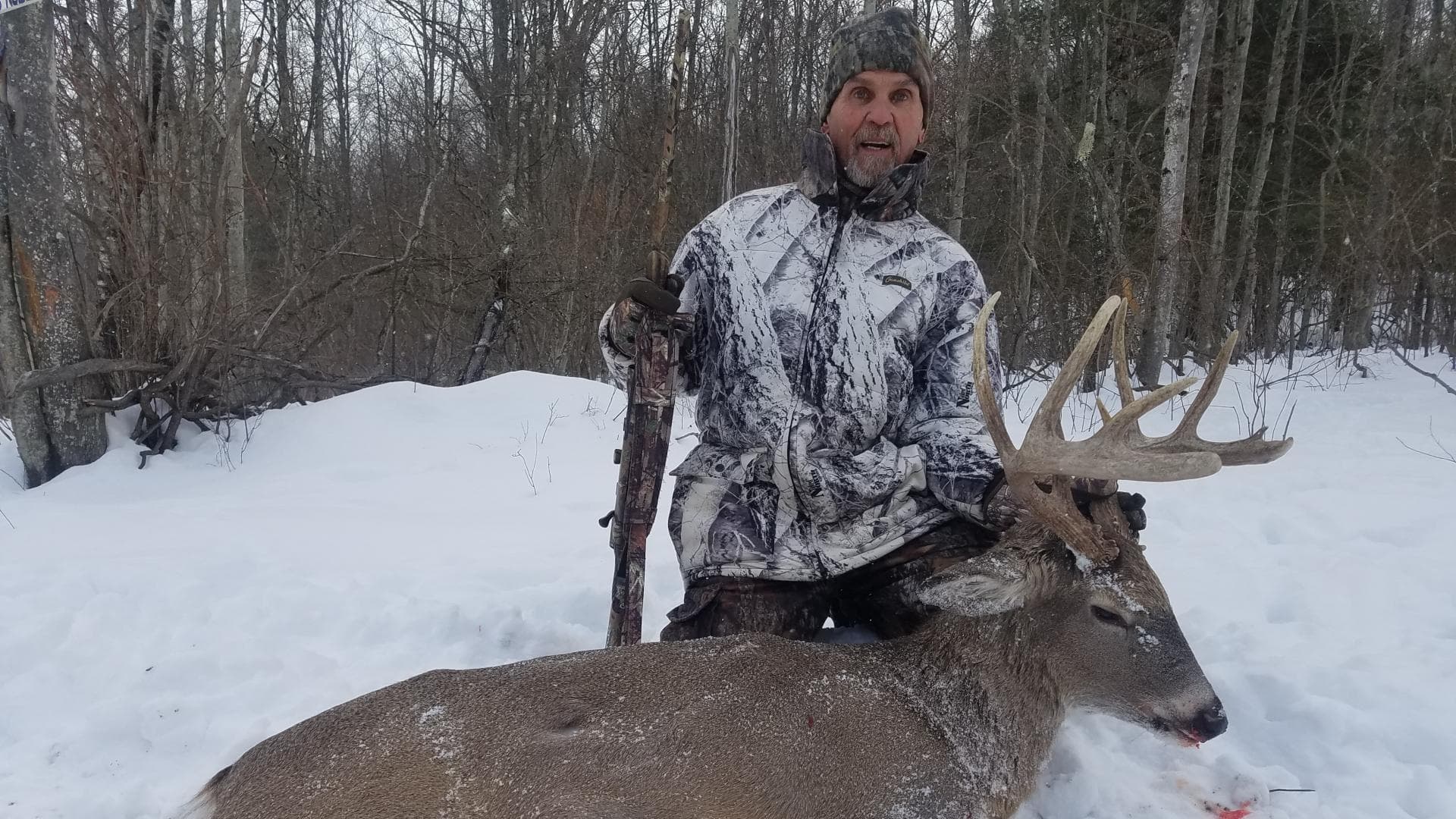 Hunter in camouflage with a large buck in the snow. Man holding a rifle, posed with the deer's large antlers. Snowy forest...