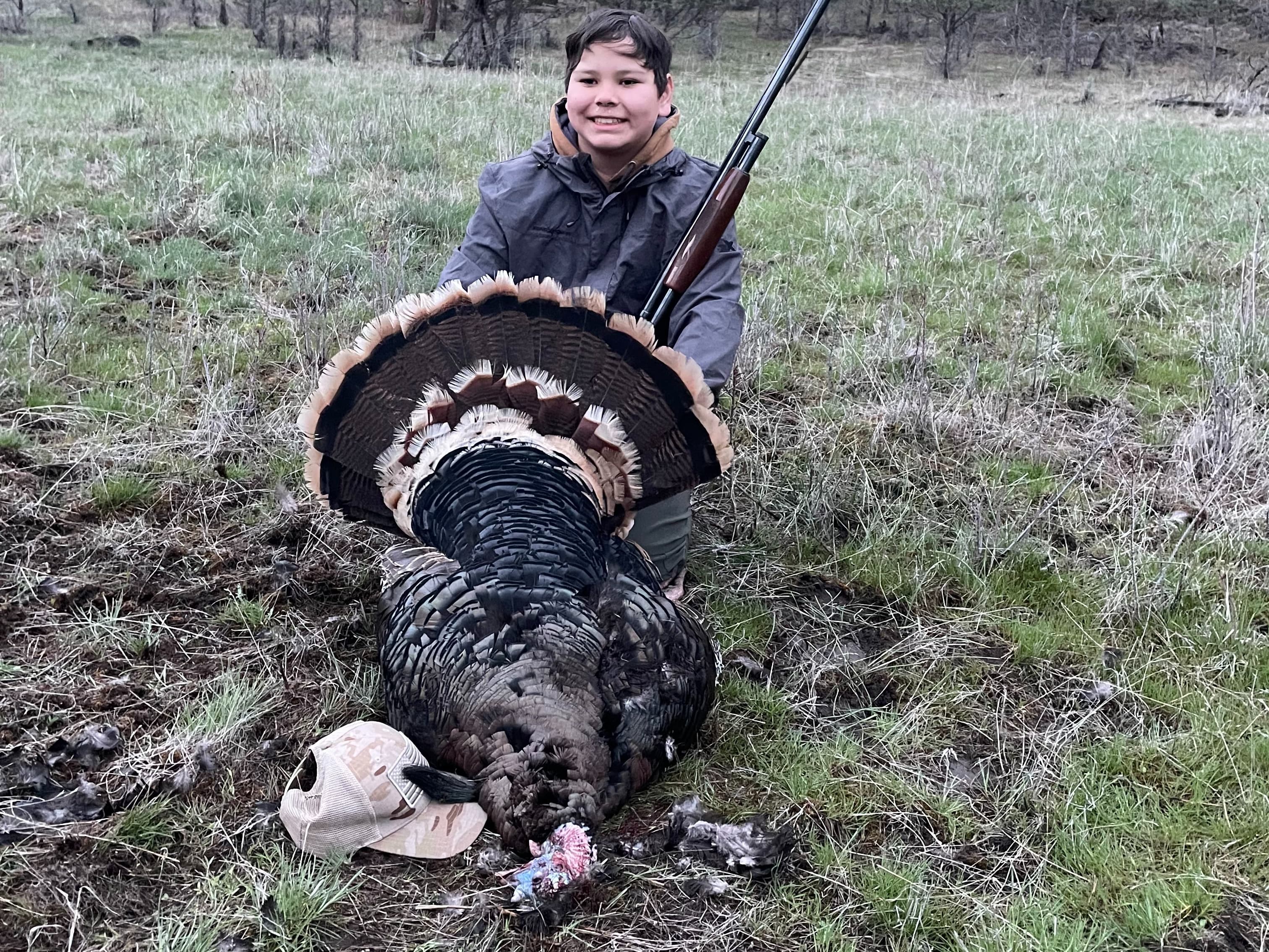 A young boy holds a dead turkey with a shotgun. The bird's fanned feathers are visible. A hat sits on the grass next to th...