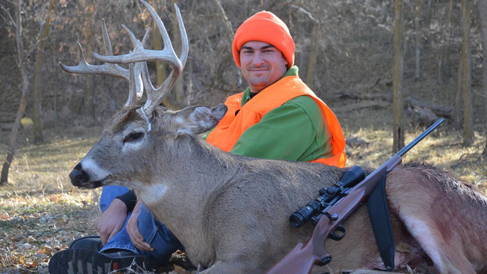 Hunter with a large buck deer. The hunter, in orange hat and vest, smiles, sitting beside the deer with large antlers. A r...