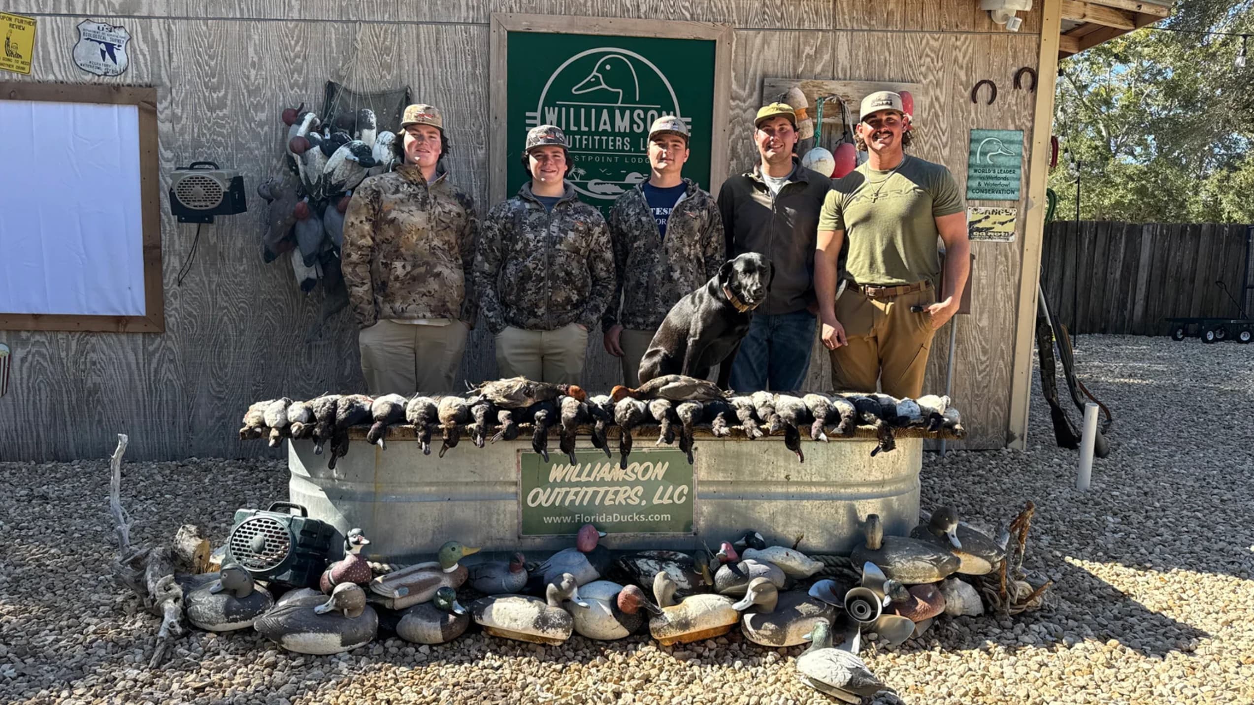 Five people pose with a black dog and a large harvest of ducks. A galvanized tub displays the ducks in front of a sign.