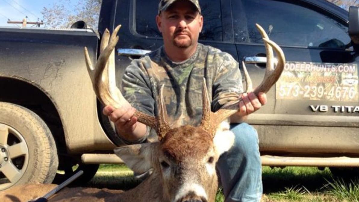 Man in camouflage holding a deer rack with prominent antlers beside a pickup truck.