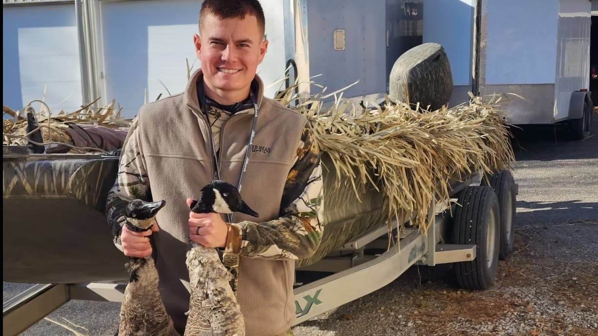 Hunter with two geese, standing next to a camouflaged duck boat on a trailer, covered in natural vegetation.