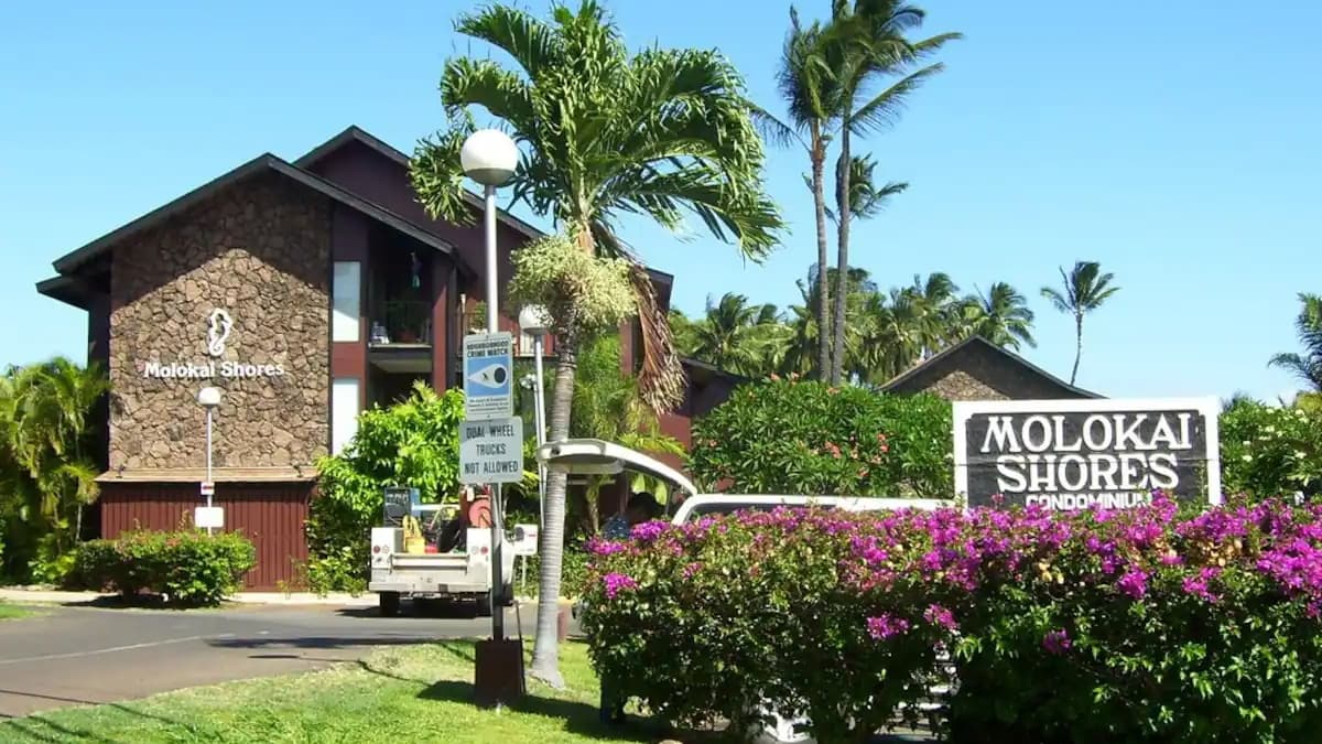 "Molokai Shores Condominium. Buildings with stone exteriors. Palm trees and flowering bushes. Sign with property name."