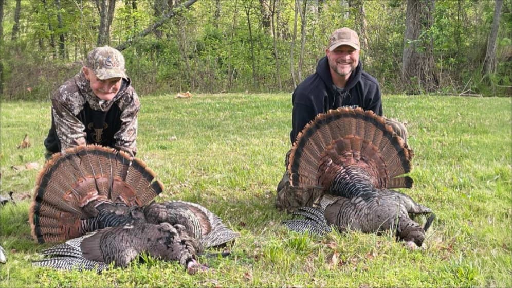 Two men kneeling with harvested turkeys, their fan feathers displayed. Forest background. Man on right smiles. Younger boy...