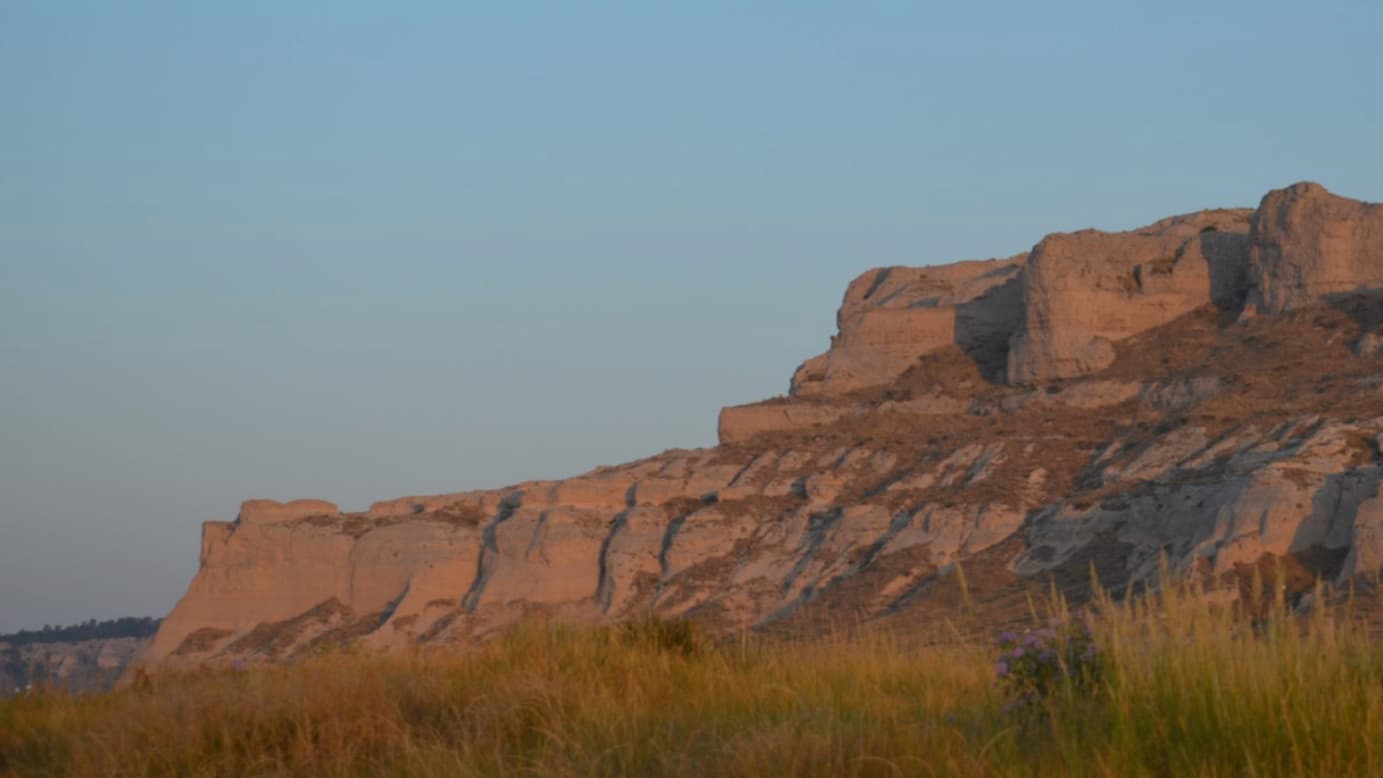 A sandstone cliff face glows in the sunset light. Tall grass and wildflowers in the foreground; pale blue sky above.