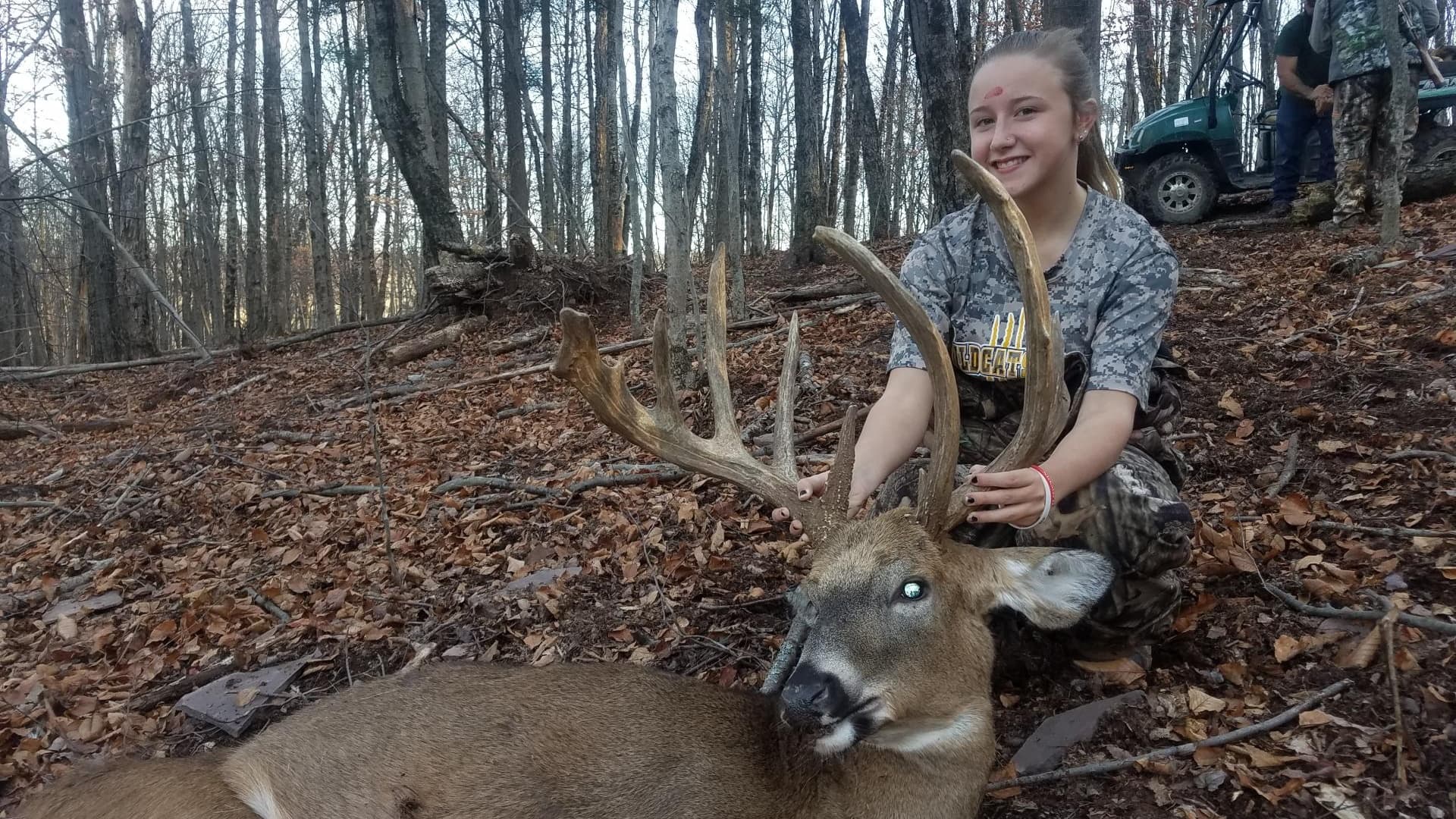 A young woman kneels beside a large buck, antlers prominent. She smiles, hands on the antlers. Autumn forest background wi...