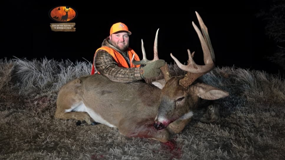 Hunter in orange vest smiles, posing with large buck. Deer has impressive antlers. Dark night setting, field grass. Blood ...