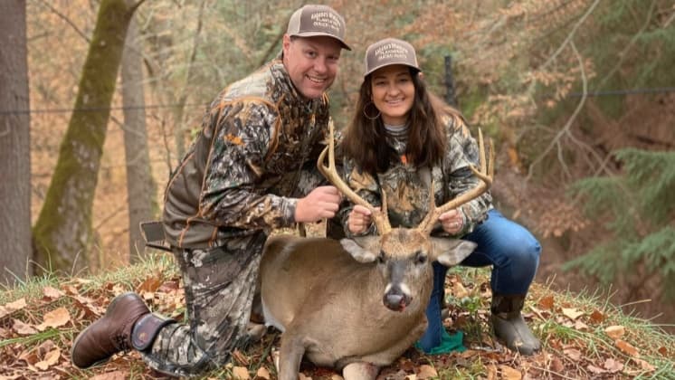 Two people in camouflage attire kneel by a large buck with antlers. They are smiling at the camera, holding the antlers ov...