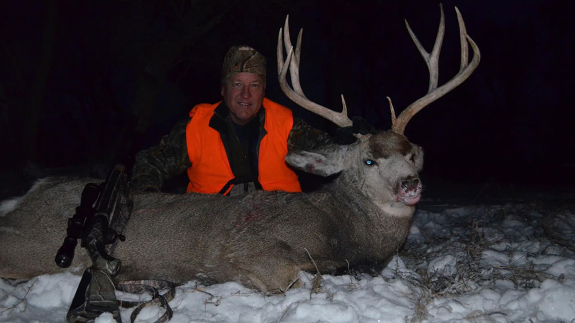 Hunter in orange vest smiles next to large buck with impressive antlers lying in snow. Rifle rests nearby. Night time scene.