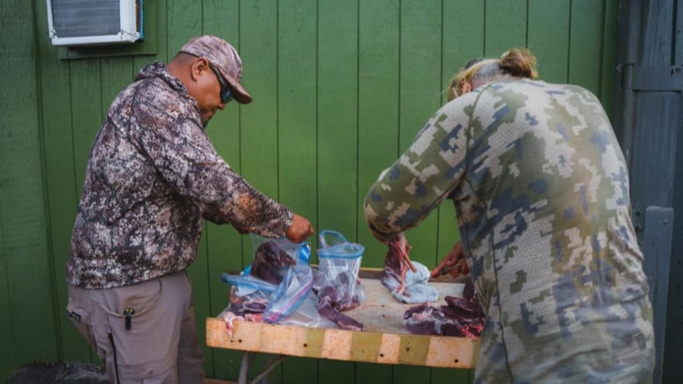 Two people in camouflage clothing are preparing meat at a table. One puts meat into bags, the other handles a piece of meat.