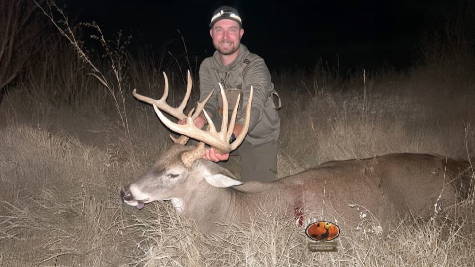 Man smiles, holding large buck antlers above the deer's body lying in tall grass. Dark outdoor setting at night.