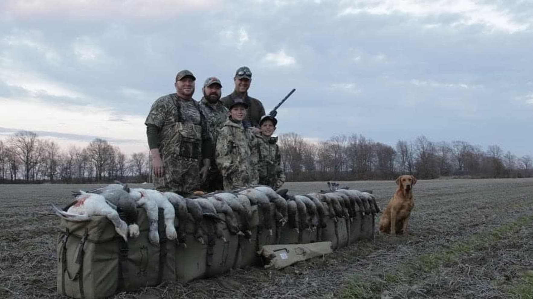 Group of five hunters pose with harvested ducks. A golden retriever sits nearby. Sky overcast.