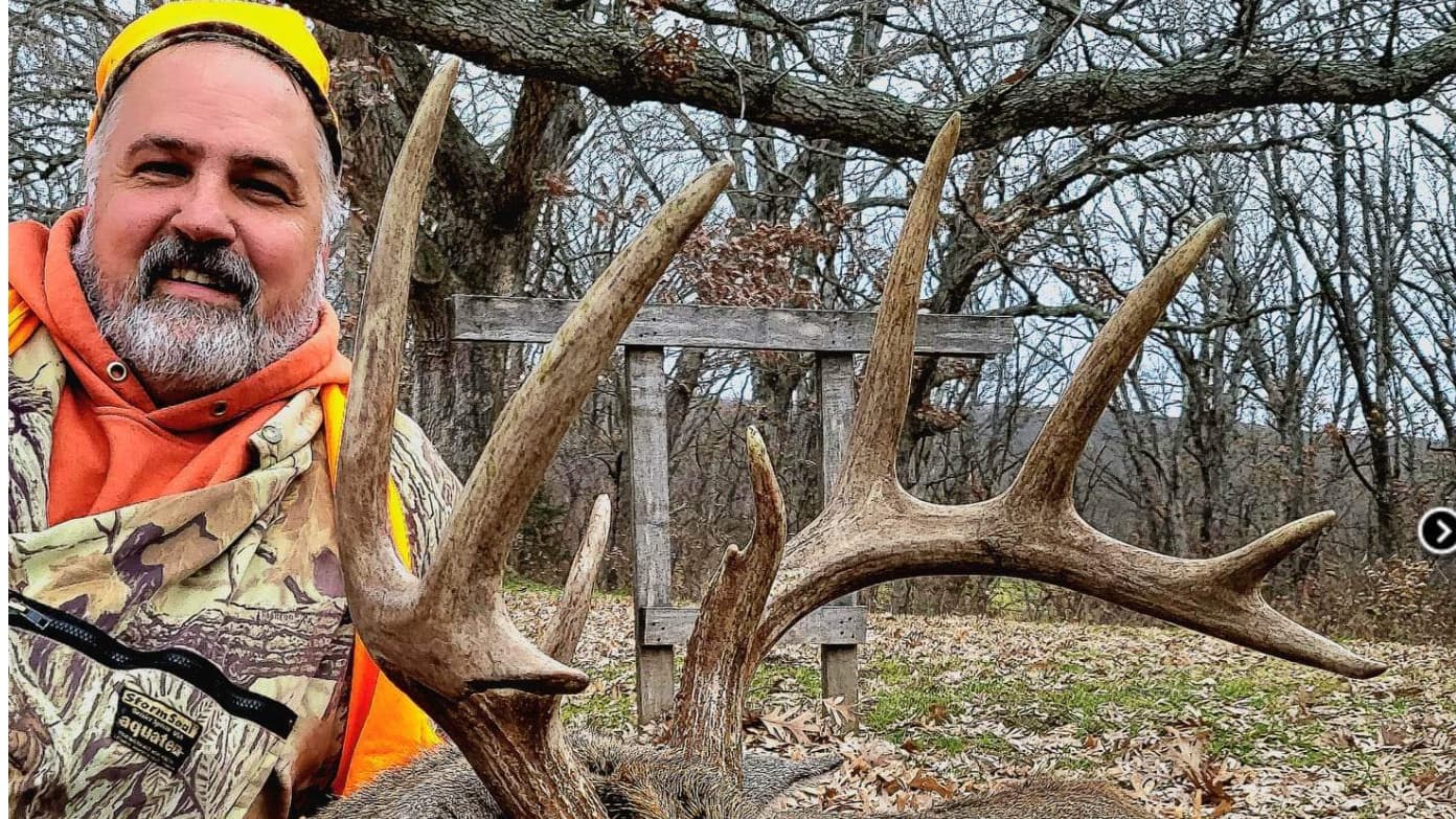 Hunter with large deer antlers and a camouflage vest in front of bare trees.