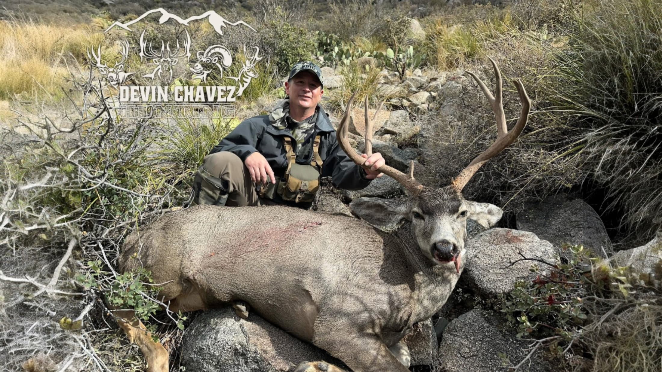 A man kneels beside a large buck with antlers. The deer lies on rocks and is surrounded by desert vegetation. The man hold...