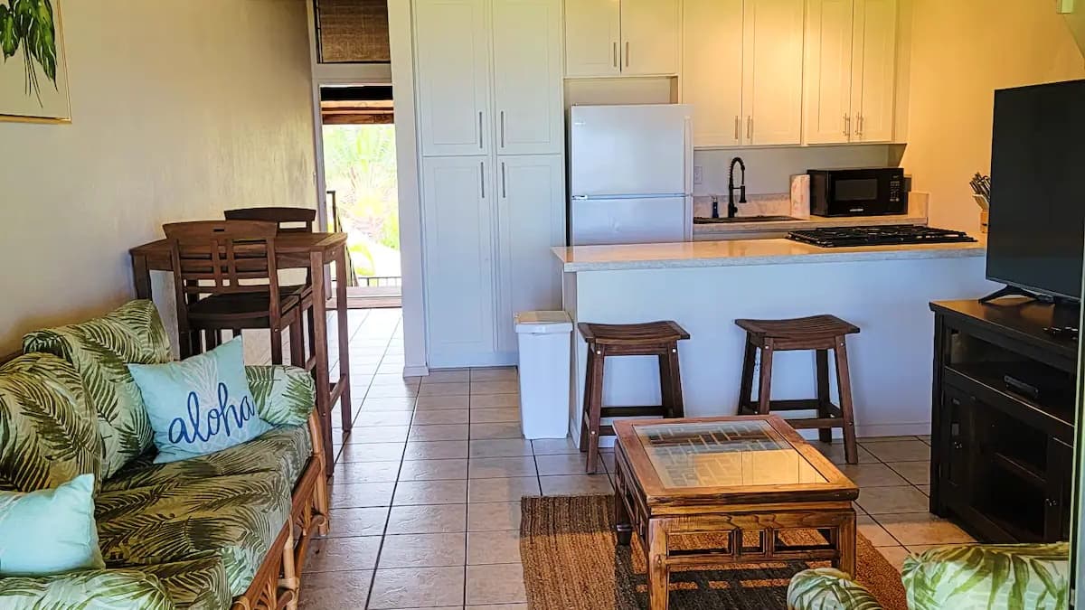 Cozy living space with kitchen. Leaf-print sofa, "Aloha" pillow, wooden table, stools, and TV. Bright white cabinets and a...