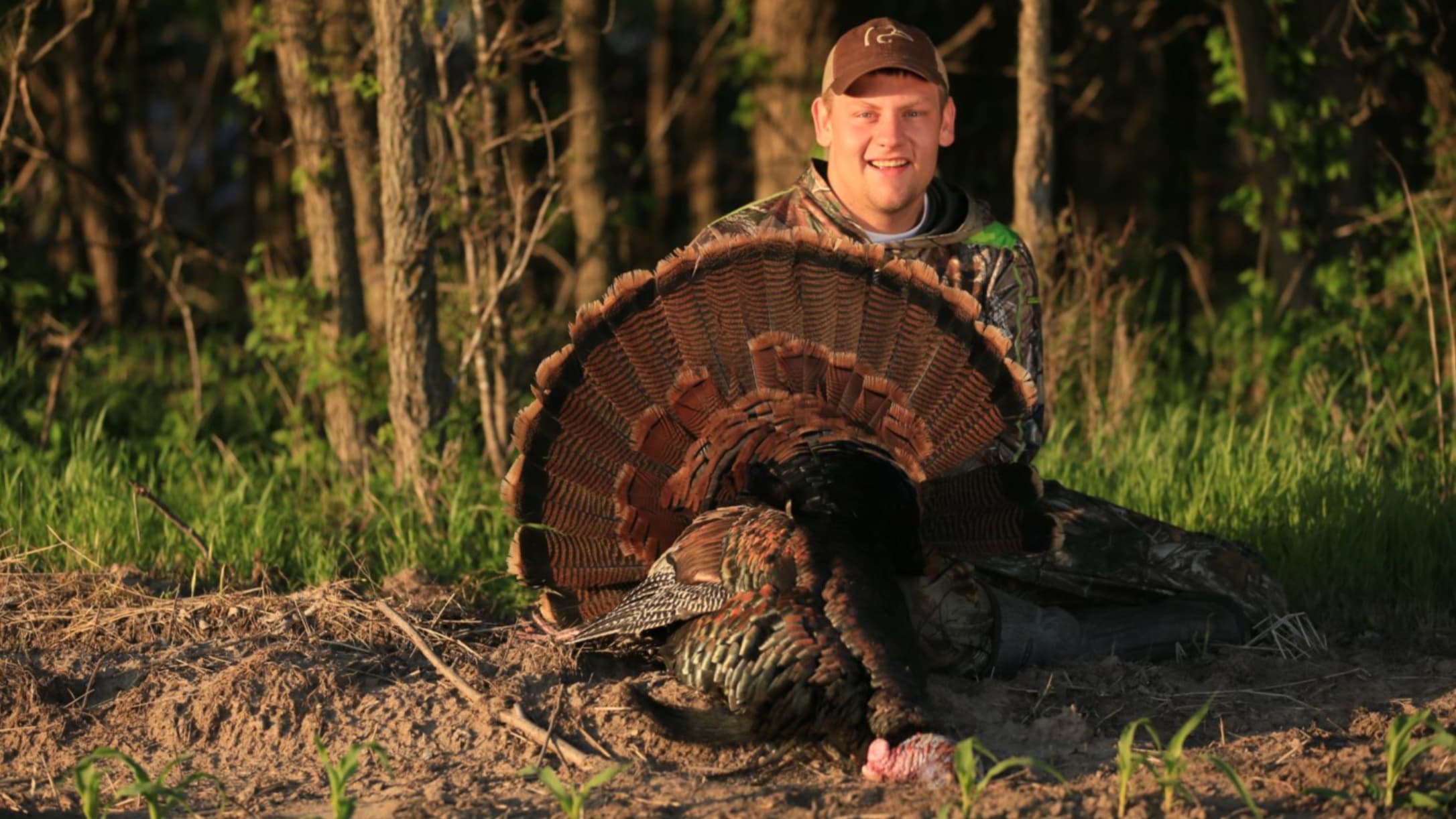 A man in camo holds a harvested turkey, its tail fanned out. He smiles at the camera, trees in the background, grass in fr...