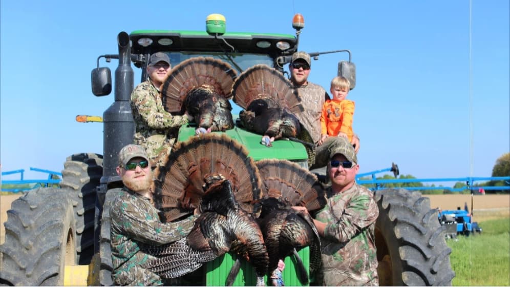 Group of hunters with turkeys posing on a John Deere tractor. Several men and a child with camouflage outfits. Large turke...