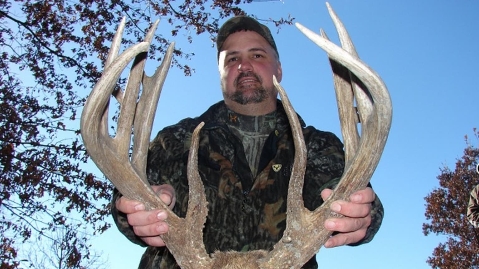 Man holding large deer antlers, looking at the camera. He wears camouflage and a cap, with a blue sky background. Bare tre...