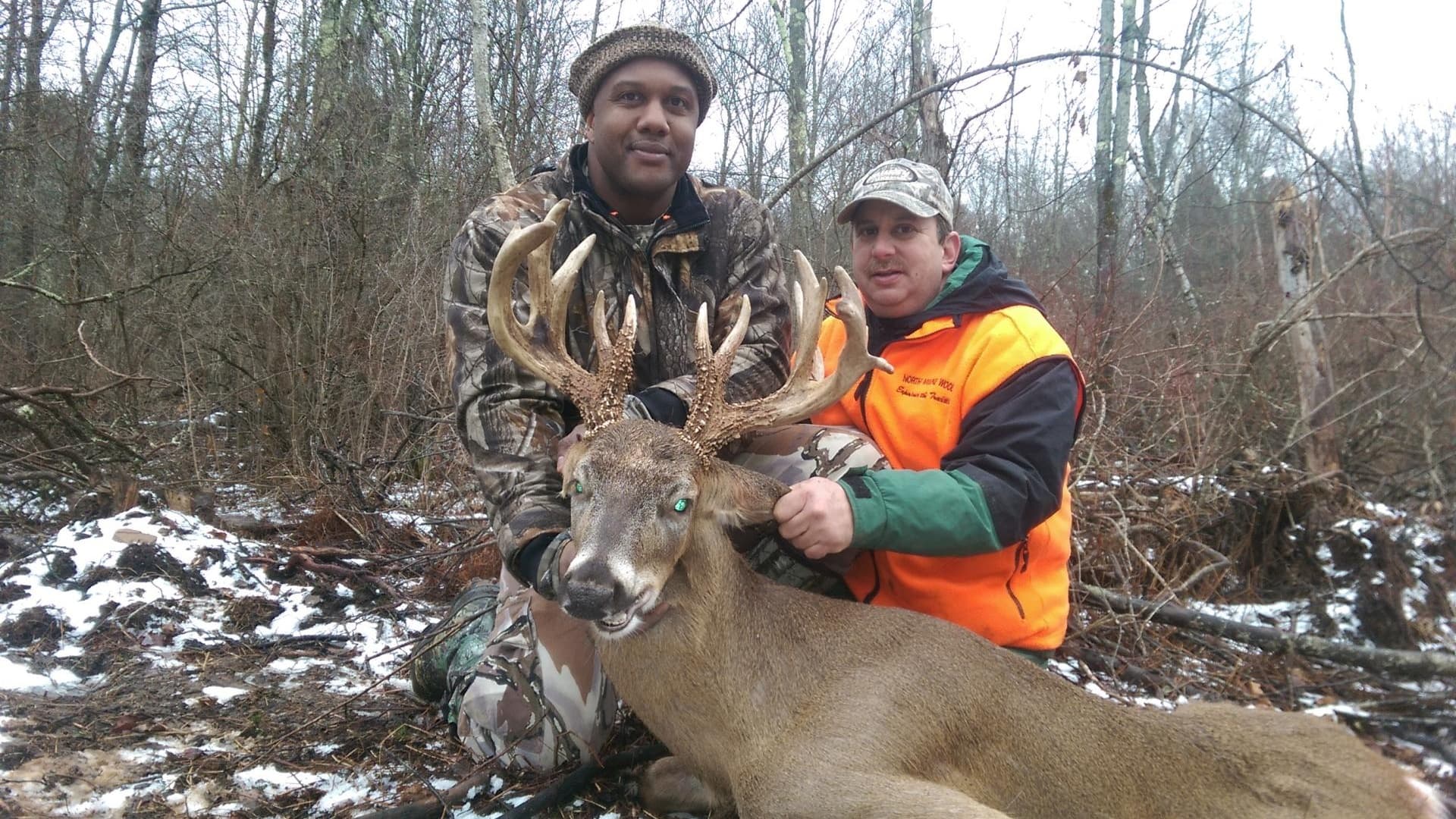 Two men pose with a large buck, holding its head. The buck has large antlers. One man wears camo, the other an orange vest...