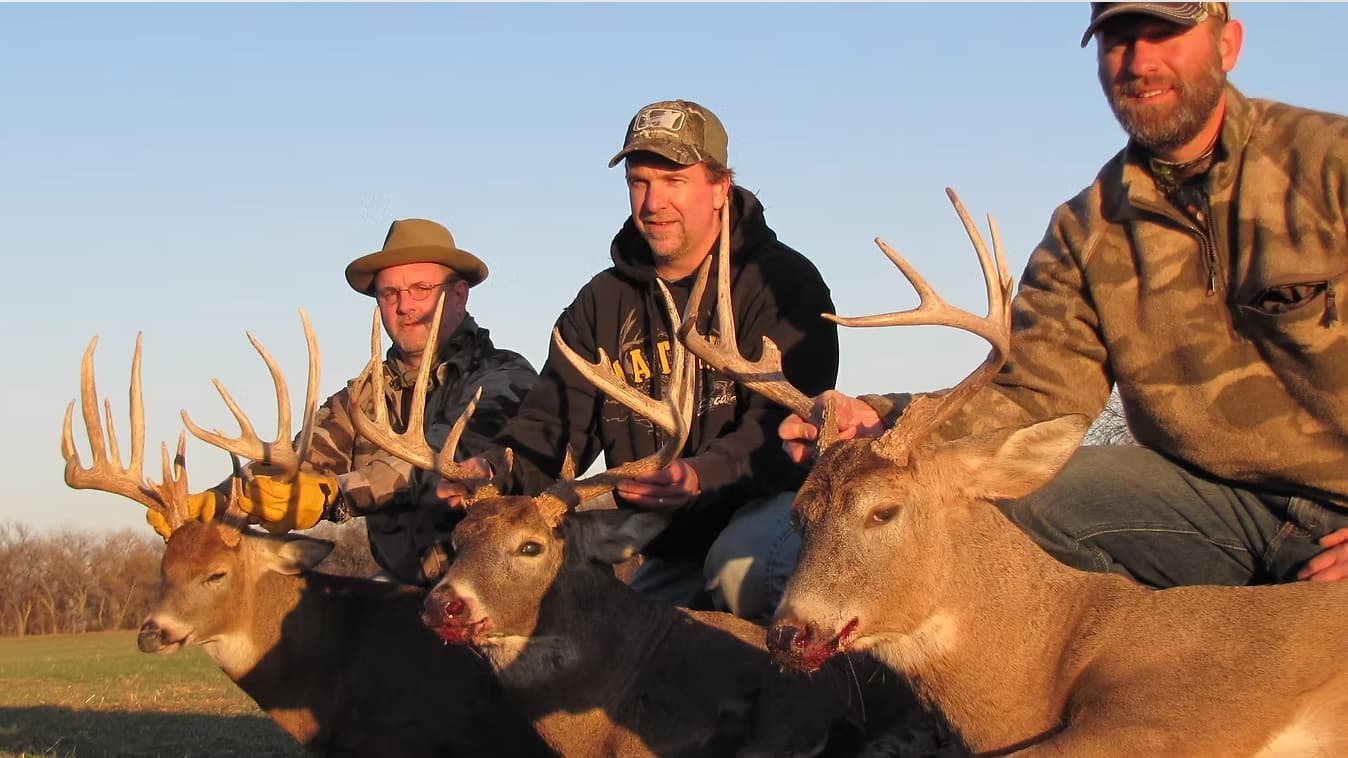 Four men with three harvested deer, holding antlers. The deer lay on grass, one with blood around its mouth. Men are smiling.