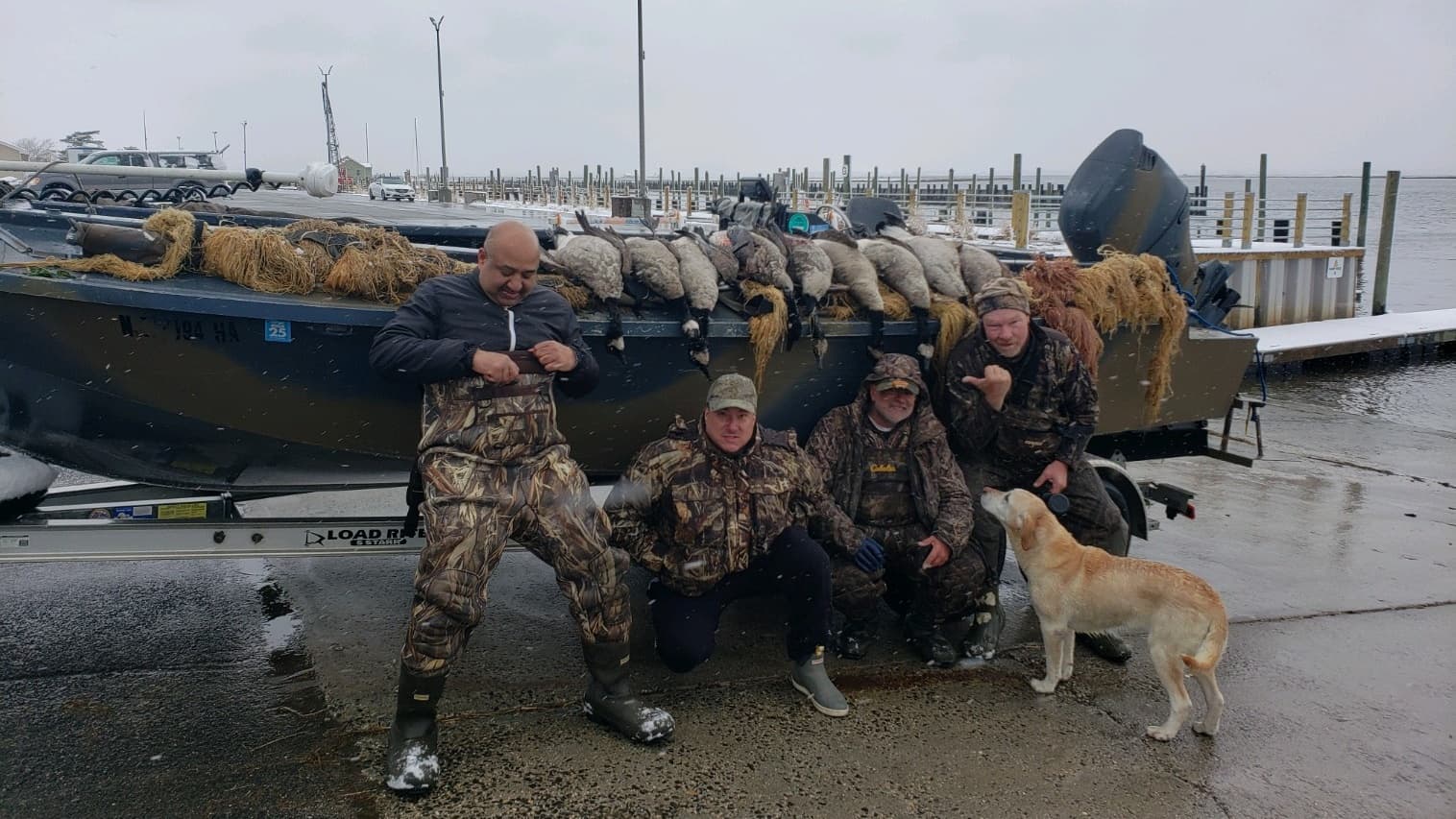 Four men in camouflage with harvested waterfowl next to a boat on a snowy dock, accompanied by a yellow lab. Boat trailer ...