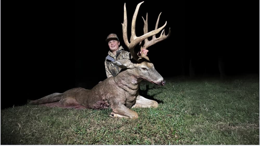 A boy smiles, standing behind a large buck with impressive antlers. The deer lies on grass. Dark background.