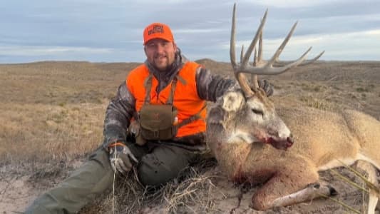 Hunter with orange vest and cap sits next to a large-antlered deer on the ground. Hunter smiles, the deer has blood on its...