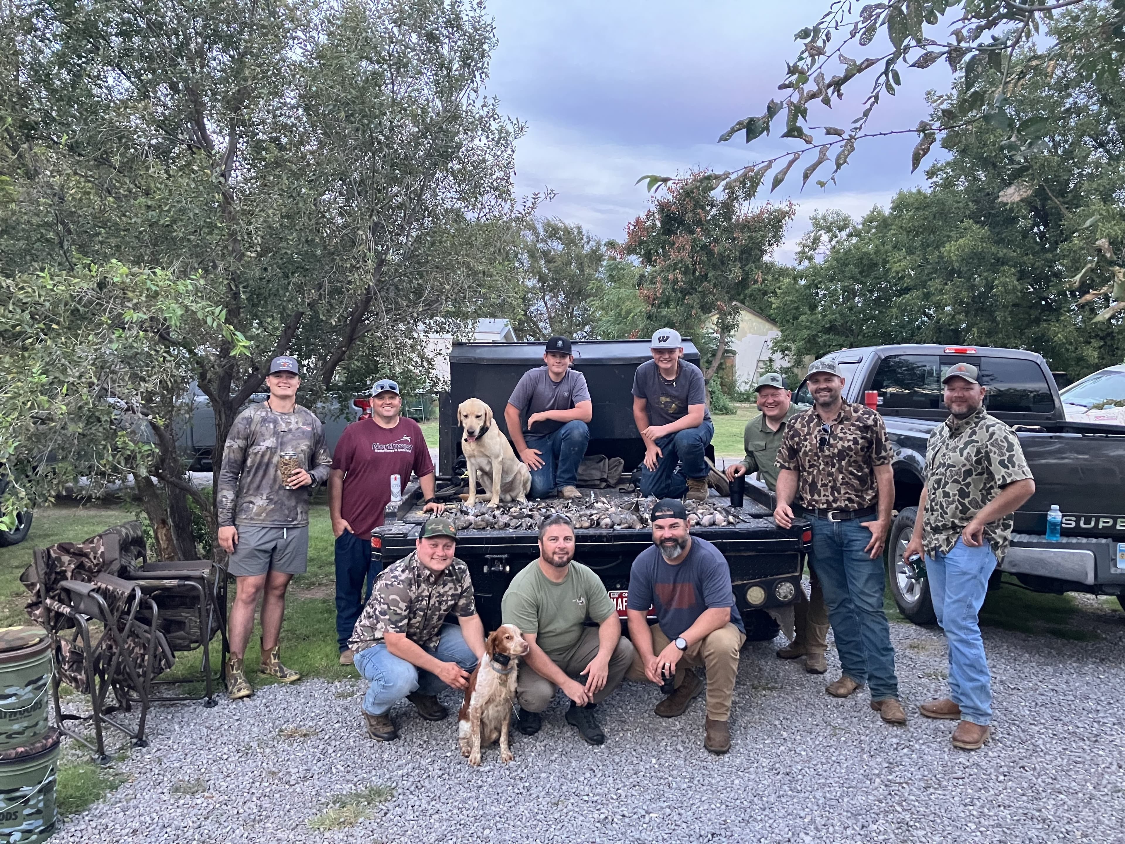 Group of men with two dogs posing by a pickup truck bed filled with hunted birds. Men wear camouflage and hats. A yellow l...