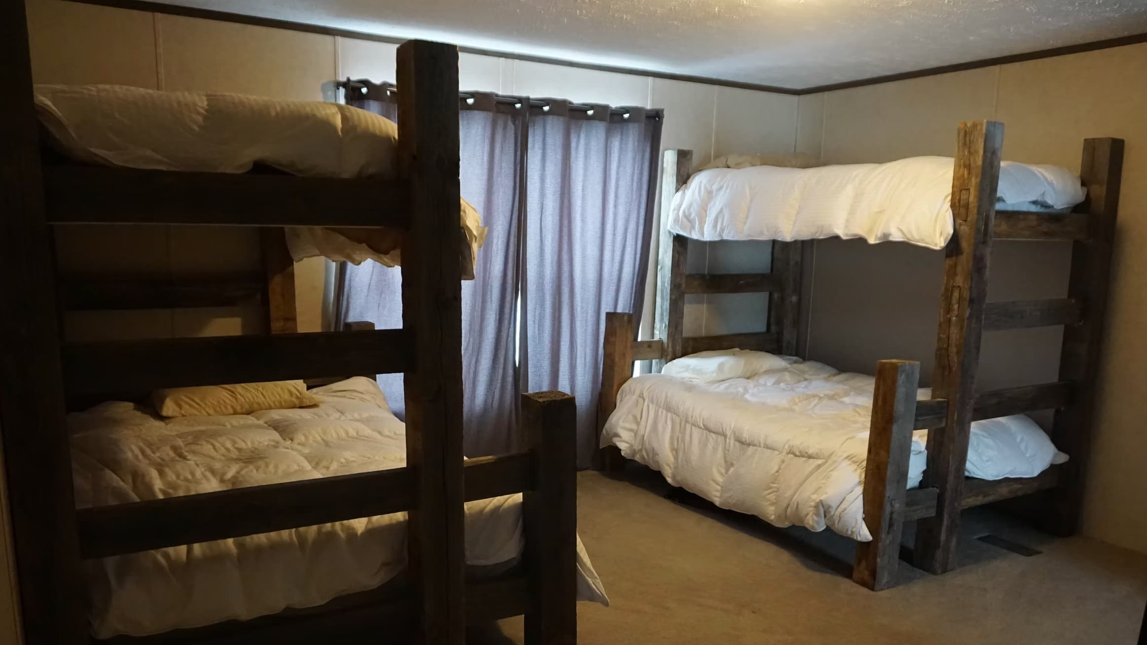 Two sets of wooden bunk beds in a room. Each bed is made with white bedding. Curtains in the background. Room lit with war...