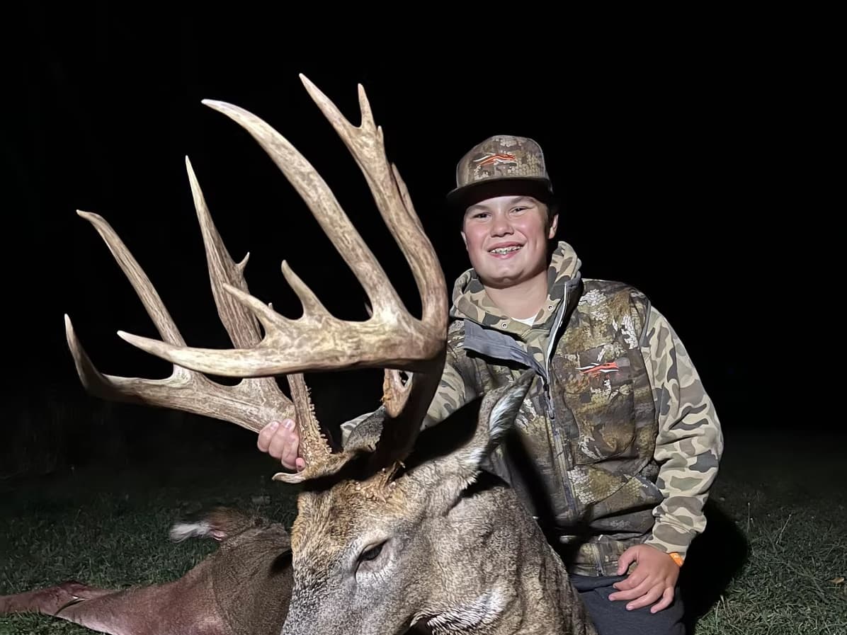 Young boy in camo, smiling, holding large deer antlers. Deer lies on ground. Dark background.