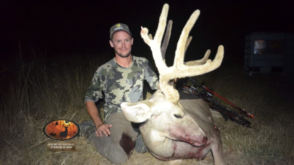 Hunter kneels beside large, white-furred buck with impressive antlers. Blood visible on buck's face. Hunter wears camo and...