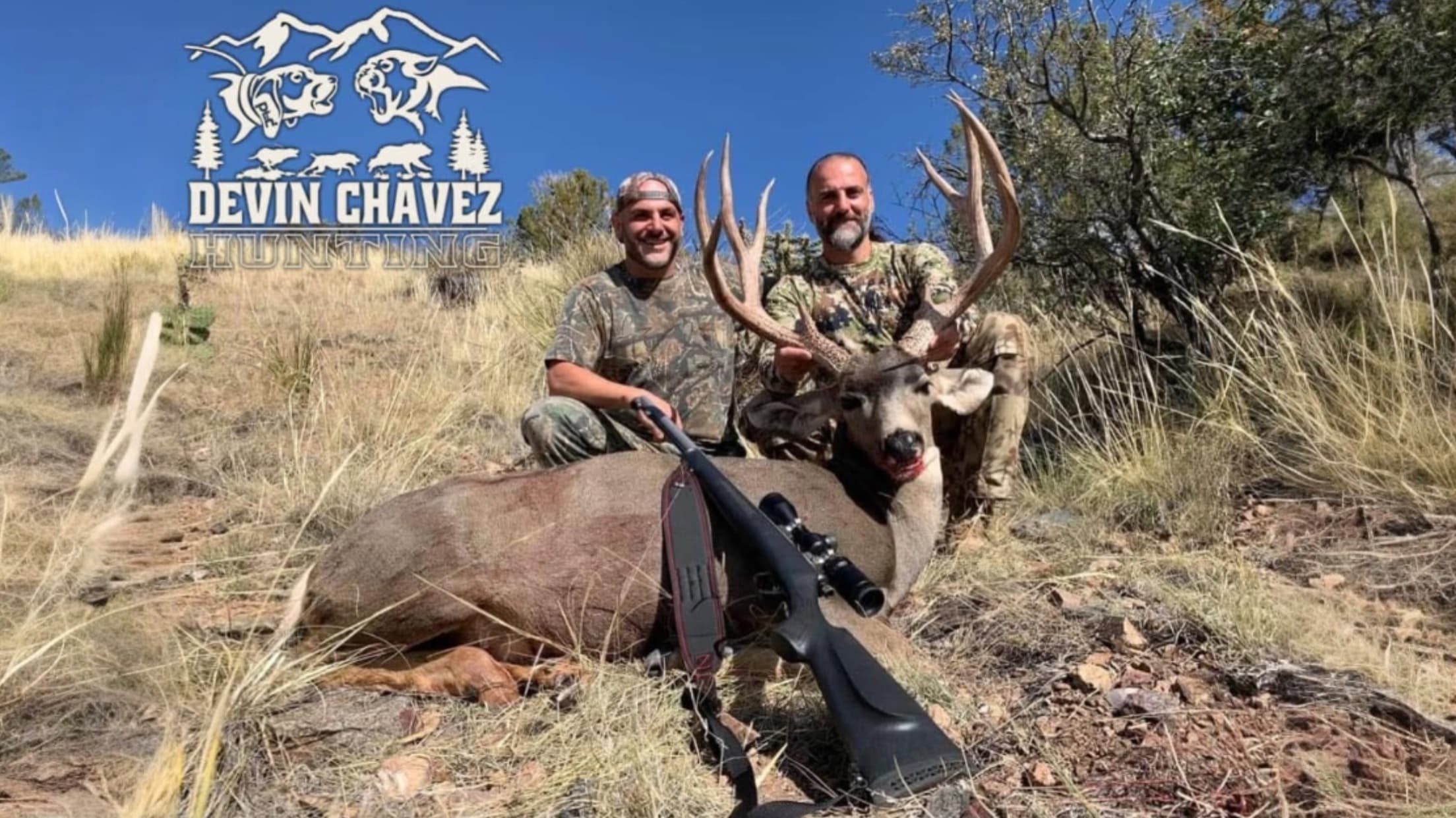 Two hunters with a large buck. One holds the antlers, other sits next to the rifle on the ground. Background: blue sky, logo.