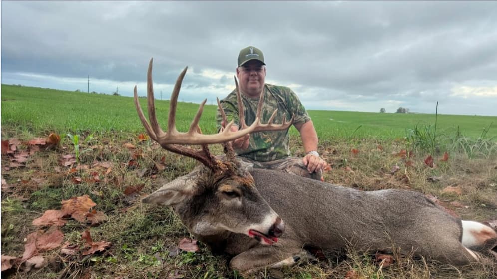 Man in camouflage poses with a large buck. Buck has large antlers and is lying on the ground. Green field and cloudy sky i...