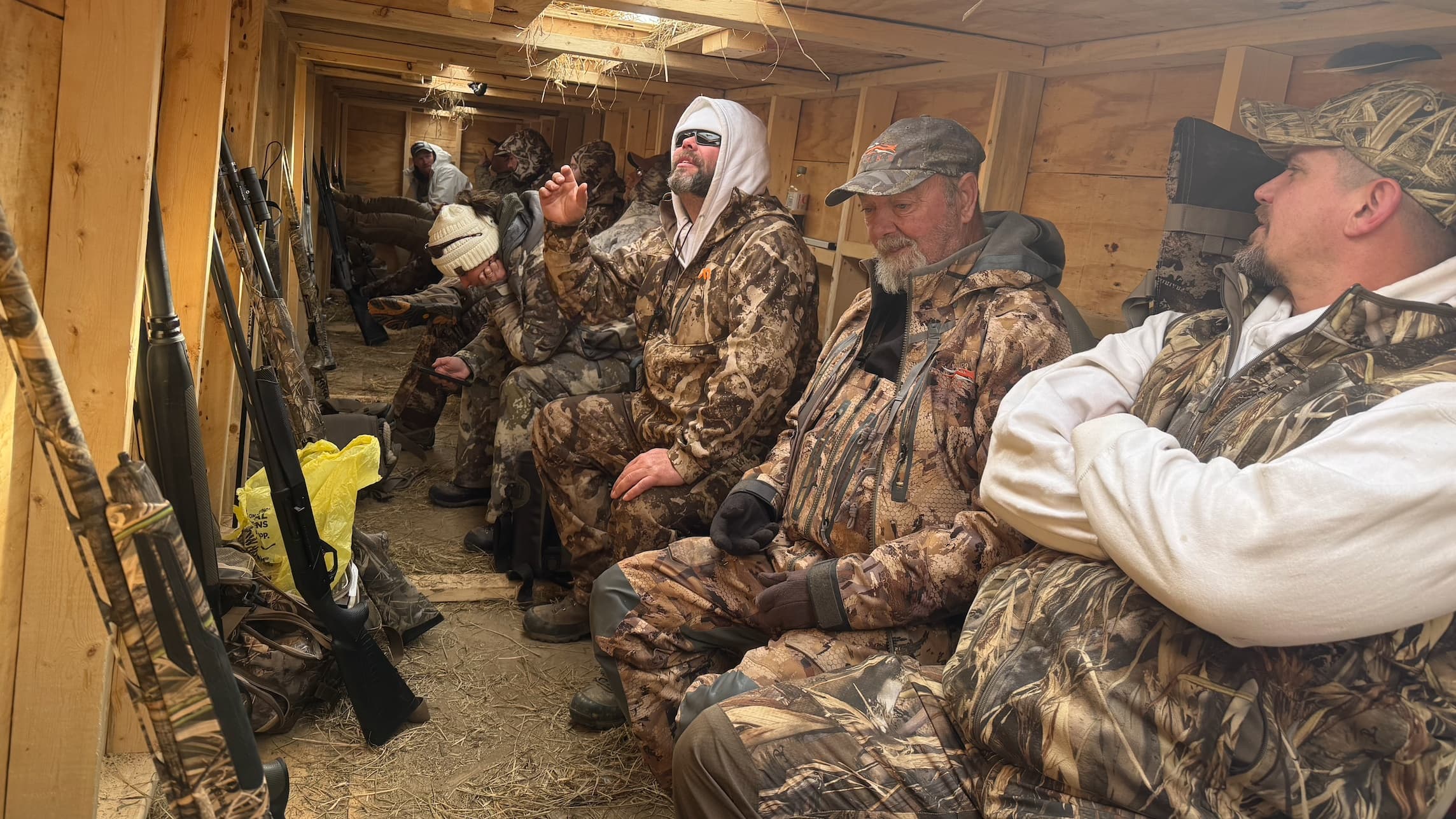 Men in camouflage attire sit inside a wooden hunting blind. Guns are leaned against the wall. Some men wear hats, sunglasses.