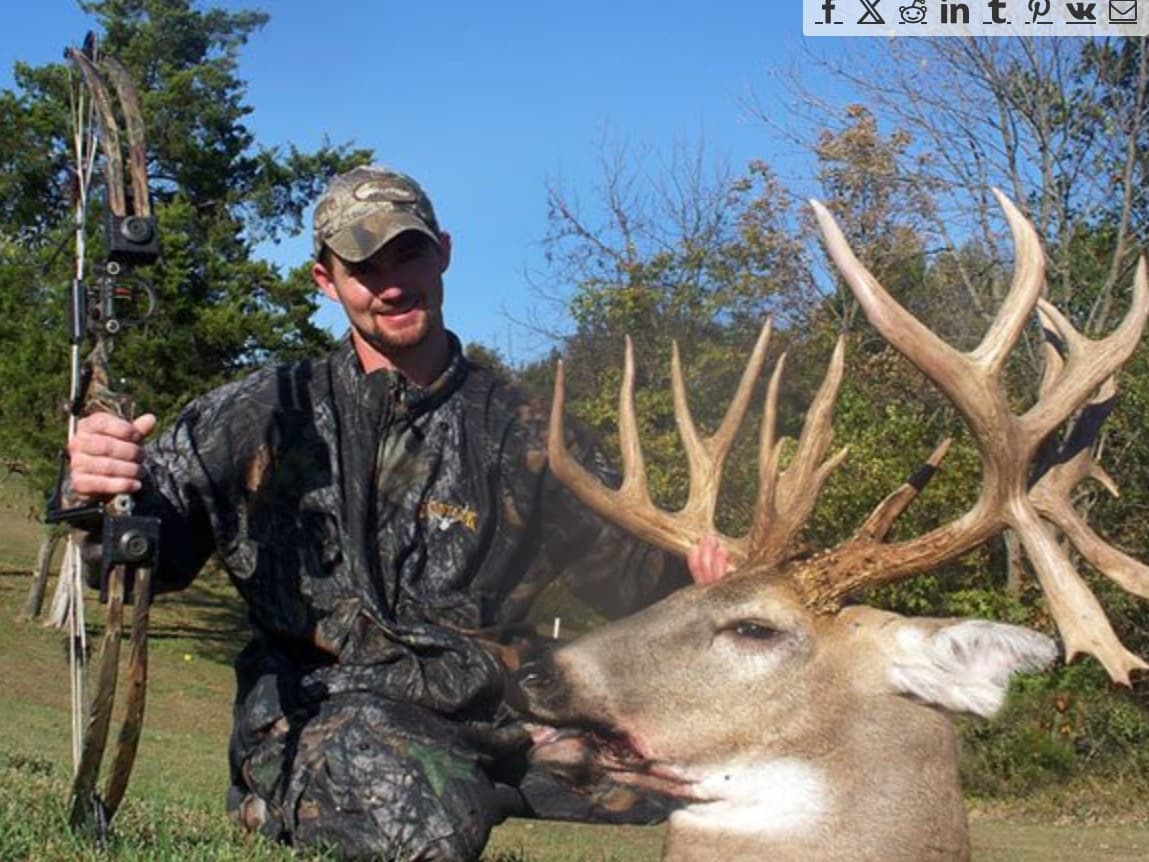 Hunter in camouflage holds a bow, posing with a large buck deer with impressive antlers.
