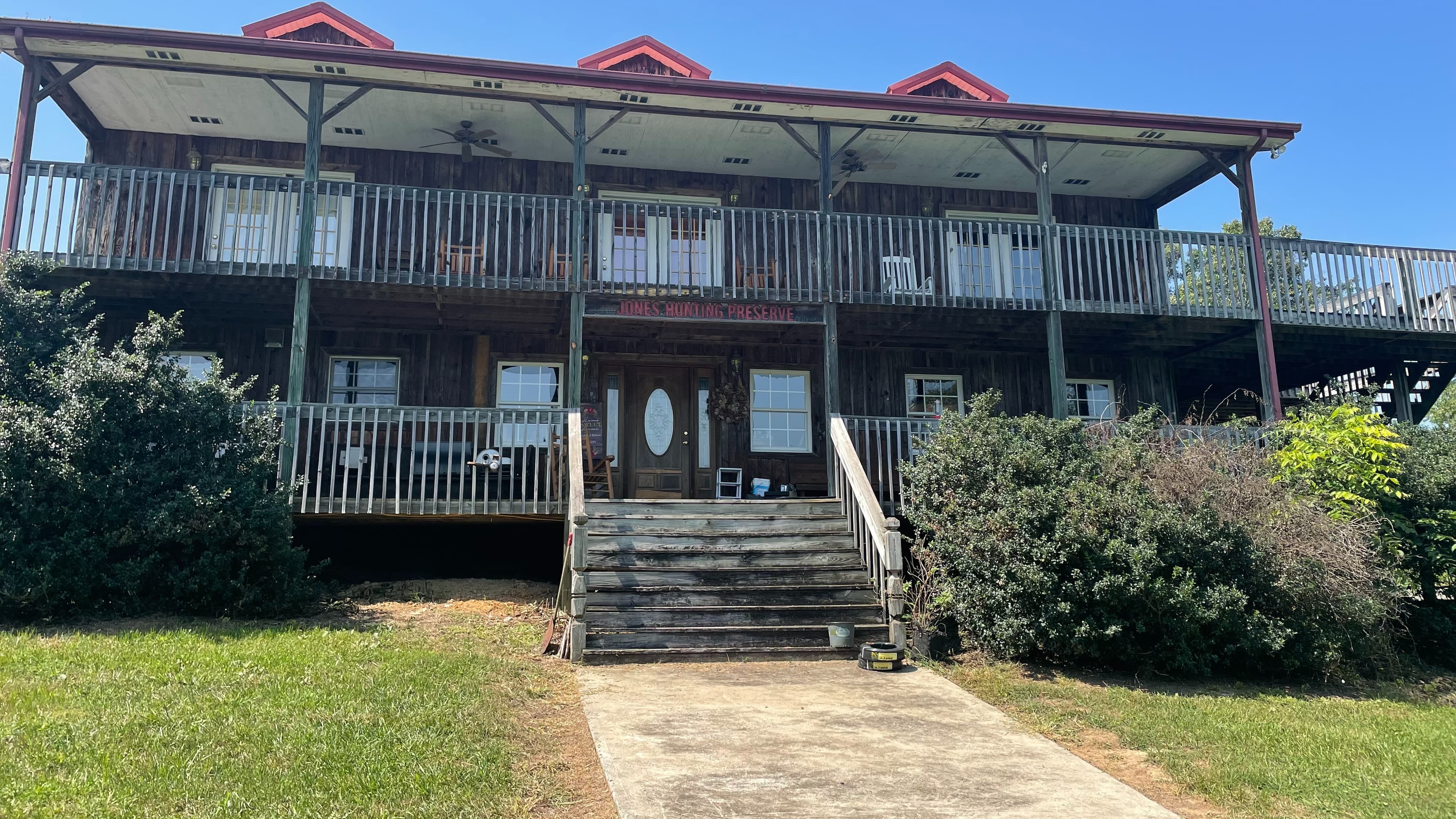 Rustic wooden lodge with two levels of balconies, stairs, and lush green surroundings under a clear blue sky.