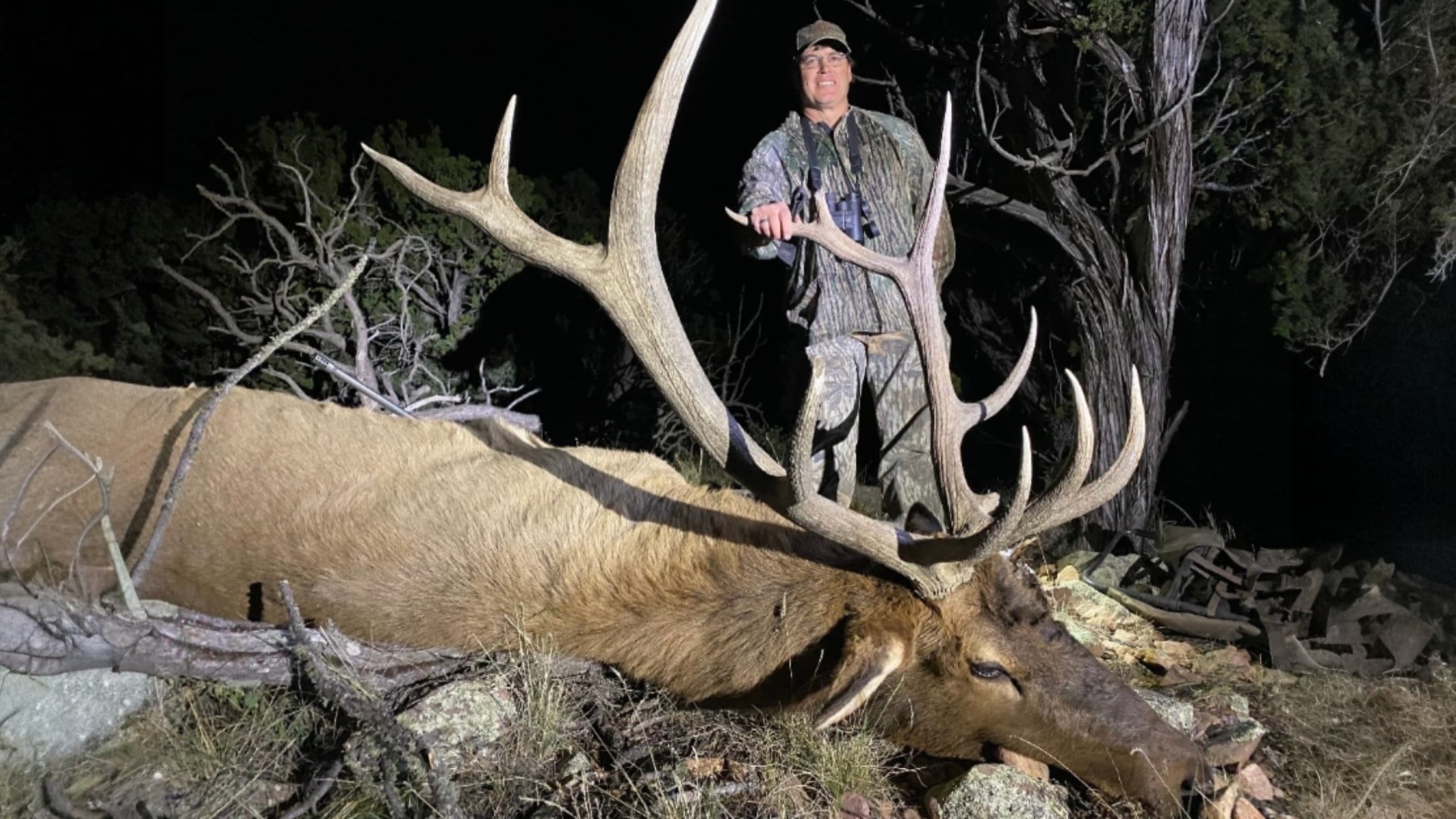 A hunter in camouflage stands proudly next to a large, dead elk with massive antlers. The animal lies on the ground, night...
