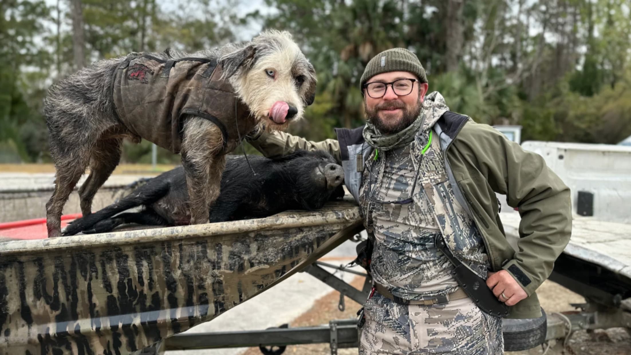 Man, dog, and dead hog in a boat. Dog in vest licks nose. Man wears camo, green jacket and hat. Hog is black. Boat has cam...