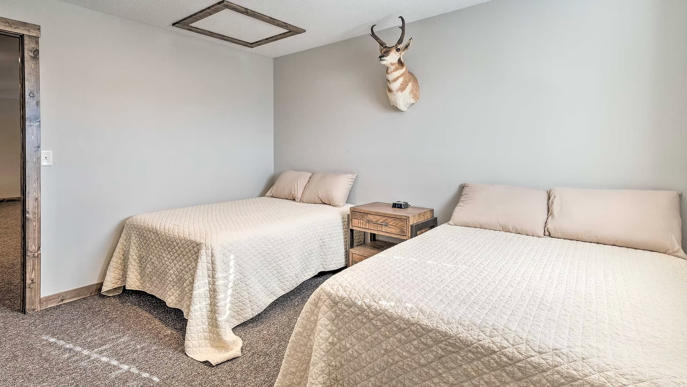 Two beds with beige quilted coverlets and pillows. Taxidermied pronghorn head on wall. Rustic bedside table between beds.