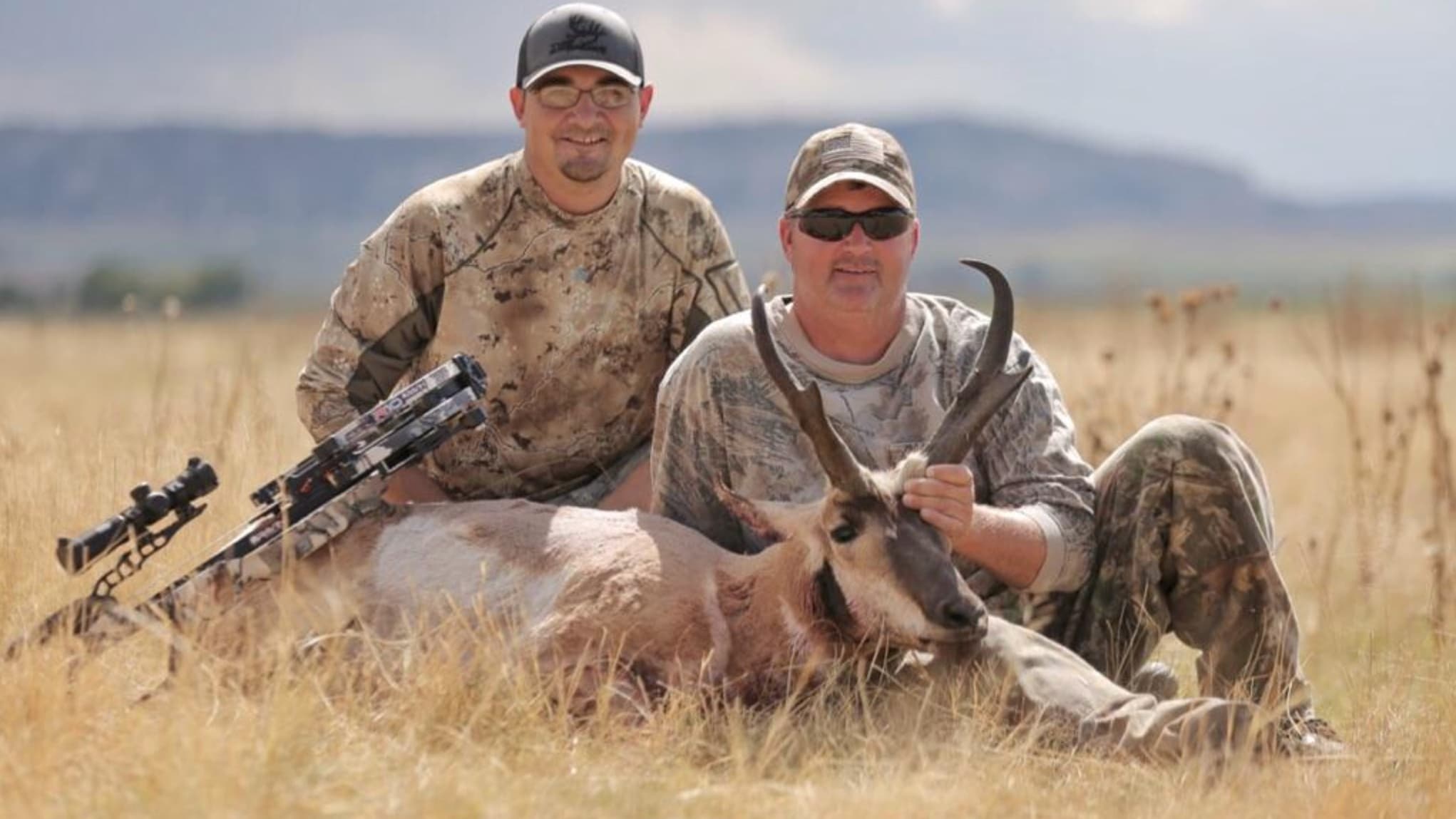 Two men posing with a dead pronghorn antelope. One man holds the antelope by its horns, while the other sits beside a cros...